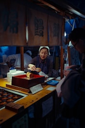 A mature woman wearing a headscarf is working at a small market stall, preparing food items for a customer. The setting appears to be a traditional food market, with various wooden containers and food trays displayed on the counter. The woman handles a large brown object, possibly food, using serving tongs. Above her, there are traditional-looking banners with Japanese characters. A sign on the stall displays a price in yen, adding to the authentic market atmosphere.