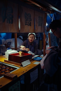 A mature woman wearing a headscarf is working at a small market stall, preparing food items for a customer. The setting appears to be a traditional food market, with various wooden containers and food trays displayed on the counter. The woman handles a large brown object, possibly food, using serving tongs. Above her, there are traditional-looking banners with Japanese characters. A sign on the stall displays a price in yen, adding to the authentic market atmosphere.
