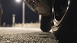 A close-up view of the tire of a vehicle, positioned in the foreground against a blurred background featuring a street lamp and pavement. The scene is captured at night, creating a moody, dimly-lit atmosphere with the focus on the shiny detail of the tire.