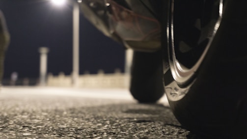 A close-up view of the tire of a vehicle, positioned in the foreground against a blurred background featuring a street lamp and pavement. The scene is captured at night, creating a moody, dimly-lit atmosphere with the focus on the shiny detail of the tire.
