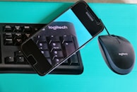 A cell phone is resting on top of a Logitech keyboard and mouse package. The phone displays an enlarged image of the keyboard, focusing on the keys with a blue background.