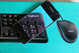 A cell phone is resting on top of a Logitech keyboard and mouse package. The phone displays an enlarged image of the keyboard, focusing on the keys with a blue background.