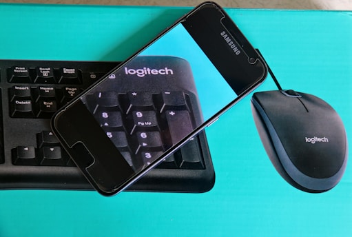 A cell phone is resting on top of a Logitech keyboard and mouse package. The phone displays an enlarged image of the keyboard, focusing on the keys with a blue background.