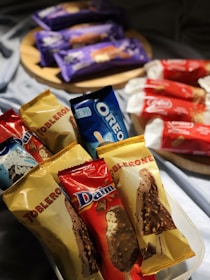 A collection of packaged ice cream bars featuring brands like Toblerone, Oreo, and Daim is displayed in a well-lit setting. The bars are arranged in an angled composition, with some lying in a plastic container in the foreground and others on a wooden tray in the background.