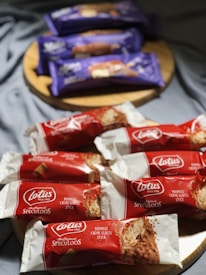 A wooden surface displays an assortment of packaged ice cream bars. In the foreground, several bars are wrapped in red and white packaging labeled 'Lotus Speculoos.' The background showcases violet and orange packaging with a slightly blurry appearance due to depth of field. The Lotus bars depict an image of the ice cream with a crumb coating.