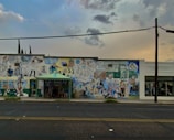 A mural on the side of a building featuring numerous colorful illustrations of people, animals, and various objects, creating a busy and vibrant scene. Next to it, there's a shop with the name 'Creations' visible on the facade. The scene is set under a cloudy sky as evening approaches, adding a dim atmosphere.