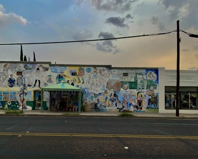 A mural on the side of a building featuring numerous colorful illustrations of people, animals, and various objects, creating a busy and vibrant scene. Next to it, there's a shop with the name 'Creations' visible on the facade. The scene is set under a cloudy sky as evening approaches, adding a dim atmosphere.