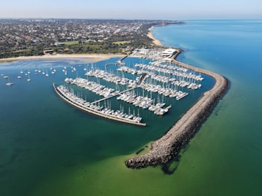 An aerial view of a marina filled with numerous boats and yachts, surrounded by a protective breakwater. The marina is situated on a coastline with a sandy beach that transitions into a densely built urban area. The calm water reflects tones of green and blue, creating a serene atmosphere.