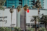 A city street scene featuring two large commercial billboards on a building facade, surrounded by palm trees. The left billboard has text, while the right one displays an image of a basketball player in action. The background comprises a modern glass building.