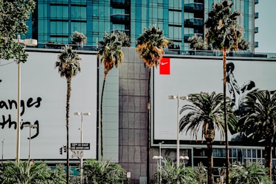 A city street scene featuring two large commercial billboards on a building facade, surrounded by palm trees. The left billboard has text, while the right one displays an image of a basketball player in action. The background comprises a modern glass building.