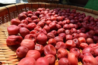 A large number of red, wrinkled plums spread out evenly on a woven bamboo mat, likely being dried in the sun. The lighting accentuates their textures and natural color variations.