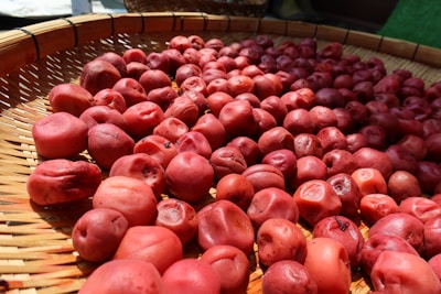 A large number of red, wrinkled plums spread out evenly on a woven bamboo mat, likely being dried in the sun. The lighting accentuates their textures and natural color variations.