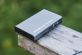 A portable external hard drive is placed on a wooden surface with a blurred green background. The device is rectangular with rounded edges and features ports on the side.