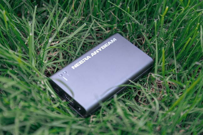 A sleek black device labeled 'NEBRA ANYBEAM' is lying on a bed of lush green grass. The device has a rectangular shape and appears to be compact and portable.