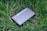 A sleek black device labeled 'NEBRA ANYBEAM' is lying on a bed of lush green grass. The device has a rectangular shape and appears to be compact and portable.