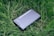 A sleek black device labeled 'NEBRA ANYBEAM' is lying on a bed of lush green grass. The device has a rectangular shape and appears to be compact and portable.