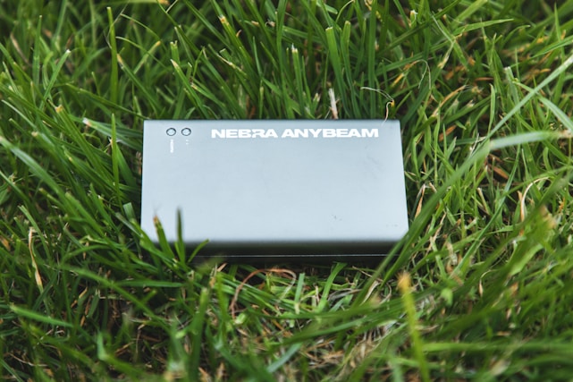 A device labeled 'NEBRA ANYBEAM' is placed on a bed of green grass. The device is rectangular and appears metallic or plastic, with two small round indicators on the top.