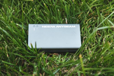 A device labeled 'NEBRA ANYBEAM' is placed on a bed of green grass. The device is rectangular and appears metallic or plastic, with two small round indicators on the top.