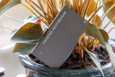 A compact, rectangular device labeled 'NEBRA ANYBEAM' is embedded in a potted plant with brown soil and large, green leaves. The device has a sleek design, featuring buttons and a port labeled 'HDMI'. The plant has thick stems and the pot is glazed with a dark color.