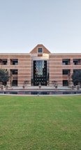 A large building with a symmetrical design featuring a central archway and numerous windows. The structure is made from red bricks and has a modern architectural style. In front of the building, there is a well-maintained lawn and a small water feature. Two trees flank the entrance, and several tables and chairs are set up near the front, suggesting a communal area.