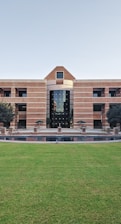 A large building with a symmetrical design featuring a central archway and numerous windows. The structure is made from red bricks and has a modern architectural style. In front of the building, there is a well-maintained lawn and a small water feature. Two trees flank the entrance, and several tables and chairs are set up near the front, suggesting a communal area.