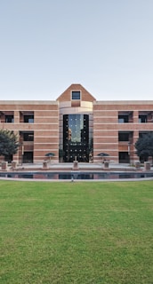 A large building with a symmetrical design featuring a central archway and numerous windows. The structure is made from red bricks and has a modern architectural style. In front of the building, there is a well-maintained lawn and a small water feature. Two trees flank the entrance, and several tables and chairs are set up near the front, suggesting a communal area.