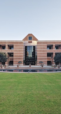 A large building with a symmetrical design featuring a central archway and numerous windows. The structure is made from red bricks and has a modern architectural style. In front of the building, there is a well-maintained lawn and a small water feature. Two trees flank the entrance, and several tables and chairs are set up near the front, suggesting a communal area.