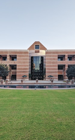 A large building with a symmetrical design featuring a central archway and numerous windows. The structure is made from red bricks and has a modern architectural style. In front of the building, there is a well-maintained lawn and a small water feature. Two trees flank the entrance, and several tables and chairs are set up near the front, suggesting a communal area.