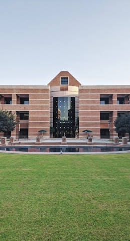 A large building with a symmetrical design featuring a central archway and numerous windows. The structure is made from red bricks and has a modern architectural style. In front of the building, there is a well-maintained lawn and a small water feature. Two trees flank the entrance, and several tables and chairs are set up near the front, suggesting a communal area.
