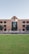 A large building with a symmetrical design featuring a central archway and numerous windows. The structure is made from red bricks and has a modern architectural style. In front of the building, there is a well-maintained lawn and a small water feature. Two trees flank the entrance, and several tables and chairs are set up near the front, suggesting a communal area.
