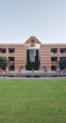 A large building with a symmetrical design featuring a central archway and numerous windows. The structure is made from red bricks and has a modern architectural style. In front of the building, there is a well-maintained lawn and a small water feature. Two trees flank the entrance, and several tables and chairs are set up near the front, suggesting a communal area.