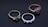 Three elegant rings are arranged on a dark reflective surface. The first ring on the left features a green gemstone set in a gold band with two smaller diamonds on each side. The middle ring is silver, displaying a row of alternating small blue sapphires and diamonds. The third ring on the right includes a pink gemstone set in a rose gold band with accompanying diamonds.