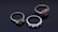 Three elegant rings are arranged on a dark reflective surface. The first ring on the left features a green gemstone set in a gold band with two smaller diamonds on each side. The middle ring is silver, displaying a row of alternating small blue sapphires and diamonds. The third ring on the right includes a pink gemstone set in a rose gold band with accompanying diamonds.