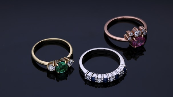 Three elegant rings are arranged on a dark reflective surface. The first ring on the left features a green gemstone set in a gold band with two smaller diamonds on each side. The middle ring is silver, displaying a row of alternating small blue sapphires and diamonds. The third ring on the right includes a pink gemstone set in a rose gold band with accompanying diamonds.