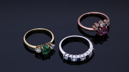 Three elegant rings are arranged on a dark reflective surface. The first ring on the left features a green gemstone set in a gold band with two smaller diamonds on each side. The middle ring is silver, displaying a row of alternating small blue sapphires and diamonds. The third ring on the right includes a pink gemstone set in a rose gold band with accompanying diamonds.