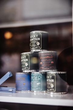 A pyramid arrangement of small, colorful, metallic tins displayed against a soft background. Each tin features distinct labels with different typography and graphics, resembling vintage or artisanal product packaging related to places like Rocky Mountain and Yosemite. A blue plastic item is visible beside the tins.