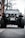 A rugged off-road vehicle with large tires is parked on a street under a clear sky. The vehicle has a distinctive grille and is equipped with additional accessories like a winch and roof rack. The setting includes bare trees and a building in the background.