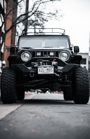 A rugged off-road vehicle with large tires is parked on a street under a clear sky. The vehicle has a distinctive grille and is equipped with additional accessories like a winch and roof rack. The setting includes bare trees and a building in the background.