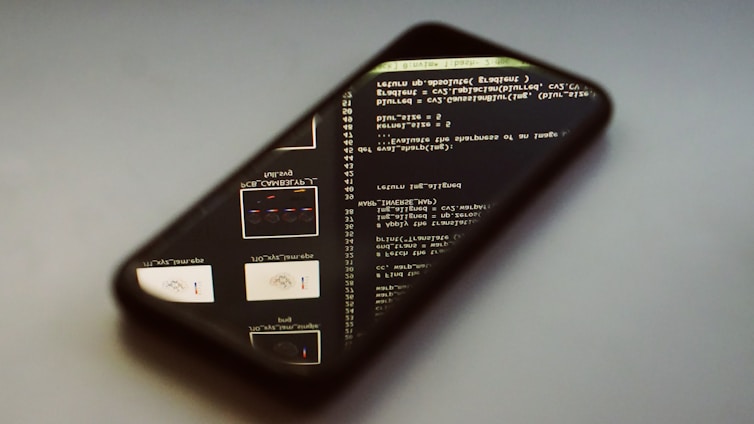 A smartphone lies on its back on a smooth surface, displaying lines of code and programming text on its screen. The screen brightness creates a soft glow, and the reflection shows the text in reverse. The phone is angled slightly to the side, partially out of focus.