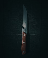A sharp kitchen knife with a wooden handle lies against a dark, textured background. The blade reflects a subtle sheen, casting faint shadows and highlighting its polished surface. Three small metal rivets secure the handle, contributing to its sturdy appearance.