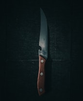 A sharp kitchen knife with a wooden handle lies against a dark, textured background. The blade reflects a subtle sheen, casting faint shadows and highlighting its polished surface. Three small metal rivets secure the handle, contributing to its sturdy appearance.