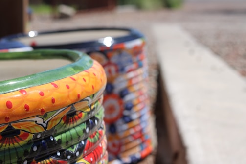 A close-up view of two colorful ceramic pots with intricate patterns. The foreground pot features vibrant hues of green, orange, and red, with detailed designs resembling fruits or abstract shapes. The background pot has a blur effect, but displays a mix of blue, orange, and white colors.