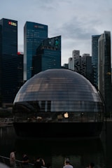 A spherical glass building sits on water, reflecting the ambient light from a city skyline filled with tall skyscrapers. The logos of various banks such as DBS and HSBC are visible on the buildings in the background. The dome structure features an Apple logo, indicating it might be an Apple store. Several people are seen standing near the structure, possibly tourists or visitors.
