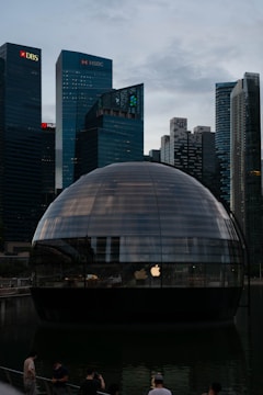 A spherical glass building sits on water, reflecting the ambient light from a city skyline filled with tall skyscrapers. The logos of various banks such as DBS and HSBC are visible on the buildings in the background. The dome structure features an Apple logo, indicating it might be an Apple store. Several people are seen standing near the structure, possibly tourists or visitors.