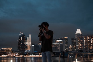A person is using a camera to take a photo during nighttime. The skyline in the background is illuminated with bright city lights, showcasing modern skyscrapers. The scene is calm with a clear, dark sky.