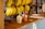 A wooden countertop displays several bunches of bananas hanging from metal hooks. In the center, there is a creamy smoothie with a straw in a cup branded with 'Fruits Cartel'. A small, colorful knitted object sits next to the smoothie, adding a playful element to the scene.