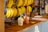 A wooden countertop displays several bunches of bananas hanging from metal hooks. In the center, there is a creamy smoothie with a straw in a cup branded with 'Fruits Cartel'. A small, colorful knitted object sits next to the smoothie, adding a playful element to the scene.