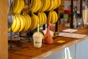 A wooden countertop displays several bunches of bananas hanging from metal hooks. In the center, there is a creamy smoothie with a straw in a cup branded with 'Fruits Cartel'. A small, colorful knitted object sits next to the smoothie, adding a playful element to the scene.