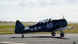 A vintage, single-engine military aircraft is stationed on a tarmac, identifiable with markings of the U.S. Navy. The aircraft has a distinctive dark blue body with the number 48 painted on the side and tail. The sky is clear with some clouds, and the grass surrounding the runway provides a vibrant contrast.