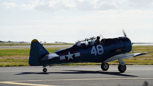 A vintage, single-engine military aircraft is stationed on a tarmac, identifiable with markings of the U.S. Navy. The aircraft has a distinctive dark blue body with the number 48 painted on the side and tail. The sky is clear with some clouds, and the grass surrounding the runway provides a vibrant contrast.